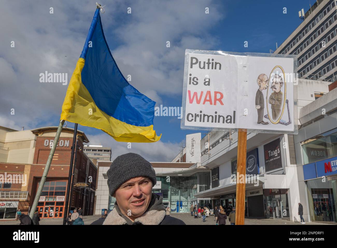 Southend on Sea, Großbritannien. Februar 2023. Ein Mann steht in Southend-on-Sea mit einer großen ukrainischen Flagge und einem Protestplakat mit der Aufschrift „Putin ist ein KRIEGSVERBRECHER“, mit einem Cartoon, der Putin zeigt, wie er Hitler im Spiegel sieht. Der Protest findet auf einem öffentlichen Platz statt, auf dem Odeon und T.K. Maxx im Hintergrund zu sehen sind. Gemeinsam in Southend halten sie ihre wöchentliche Mahnwache in der High Street ab, um Unterstützung zu zeigen und die anhaltende Situation in der Ukraine hervorzuheben. Penelope Barritt/Alamy Live News Stockfoto
