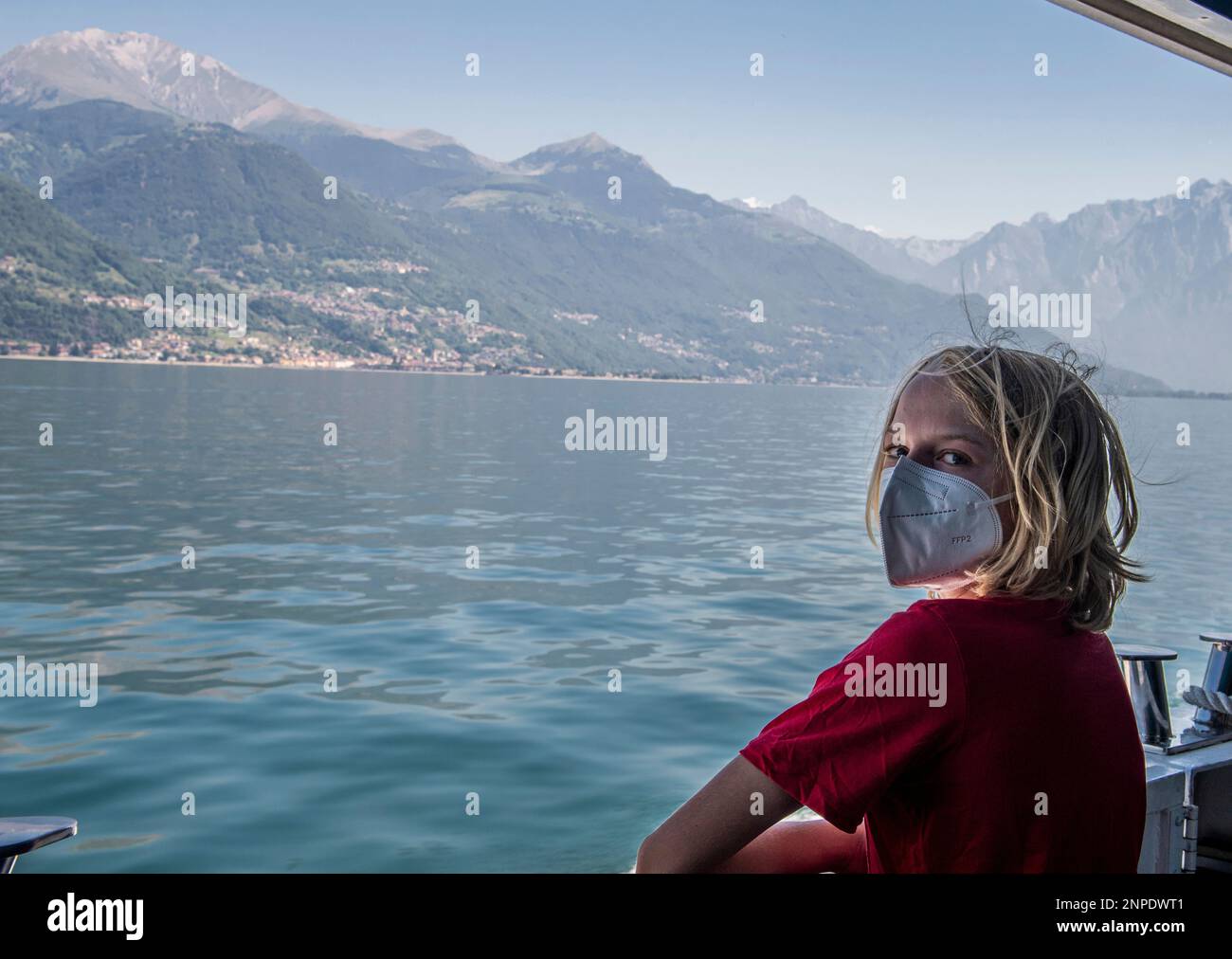 Ein Junge mit blonden Haaren und einem roten Oberteil trägt eine Gesichtsmaske in Krankenhausqualität und schaut direkt auf die Kamera, während er an Bord einer Fähre auf dem Comer See ist. Stockfoto