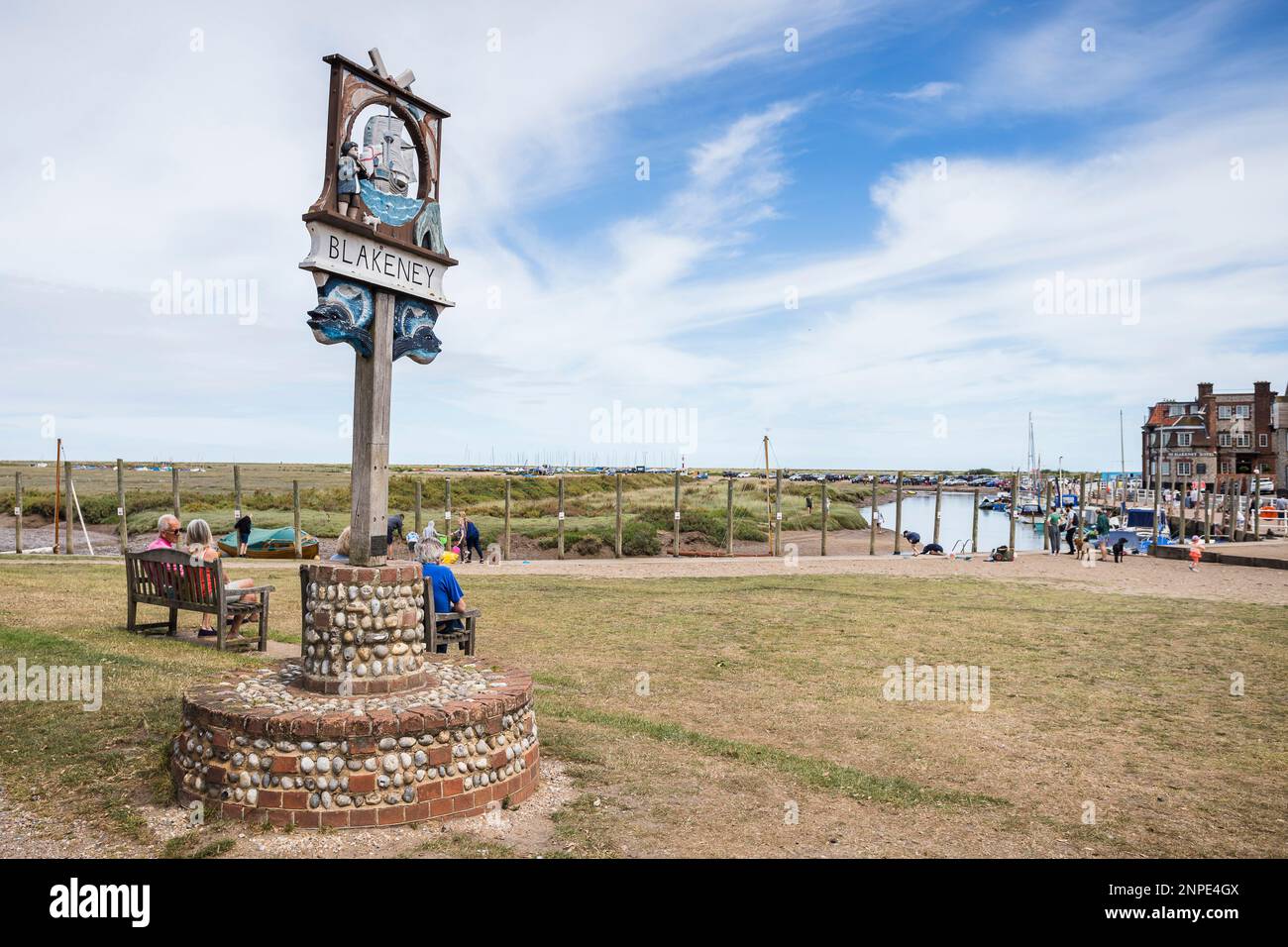 Ein Schild zum Dorf Blakeney ist neben den Bänken abgebildet, die von Urlaubern an der Nordnorfolkküste besetzt sind. Stockfoto