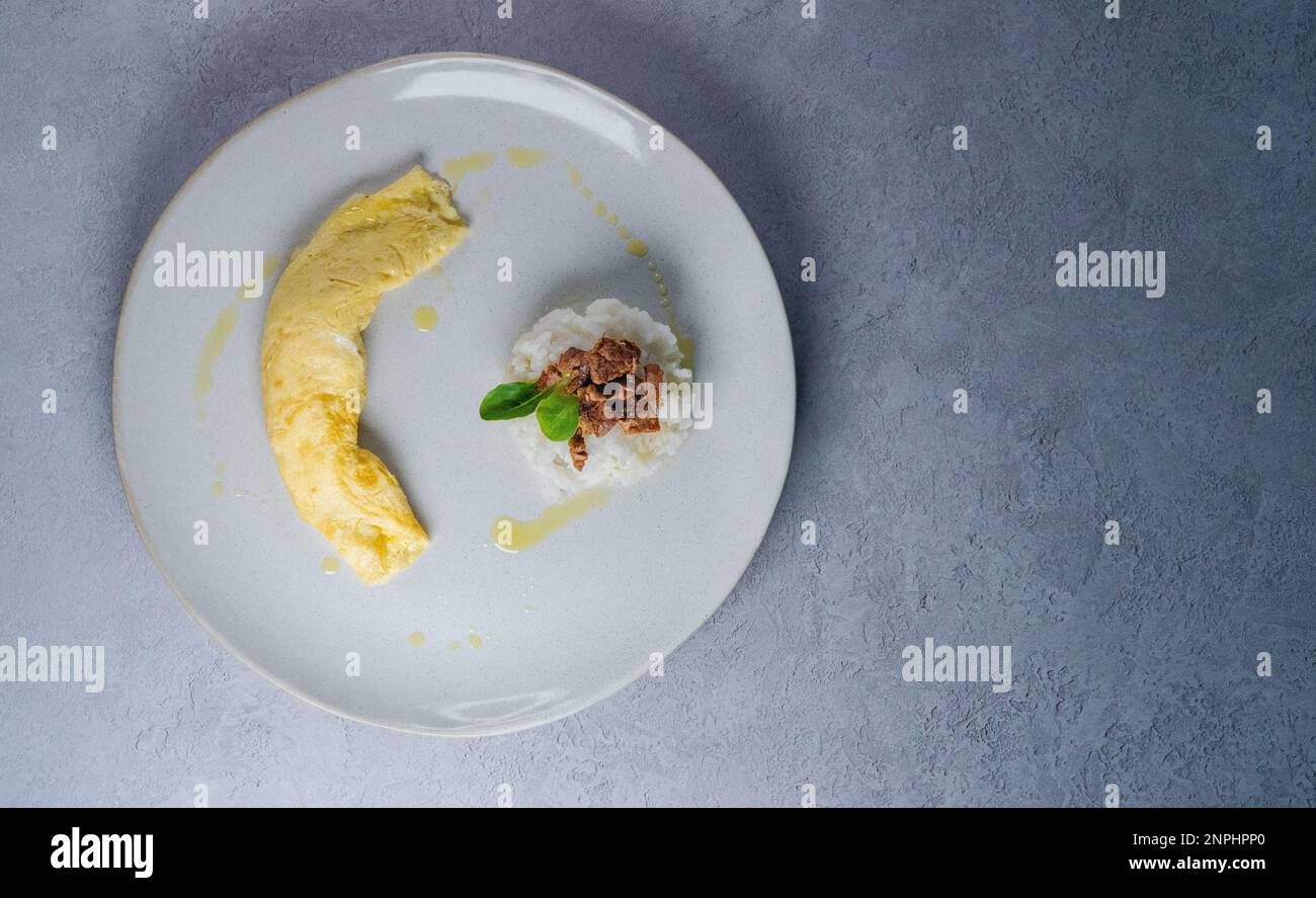 Omelette auf dem Teller mit Reis und Rindfleisch. Sprühen Sie Olivenöl und grünen Mikrosalat auf einer hellblauen Betontheke. Stockfoto