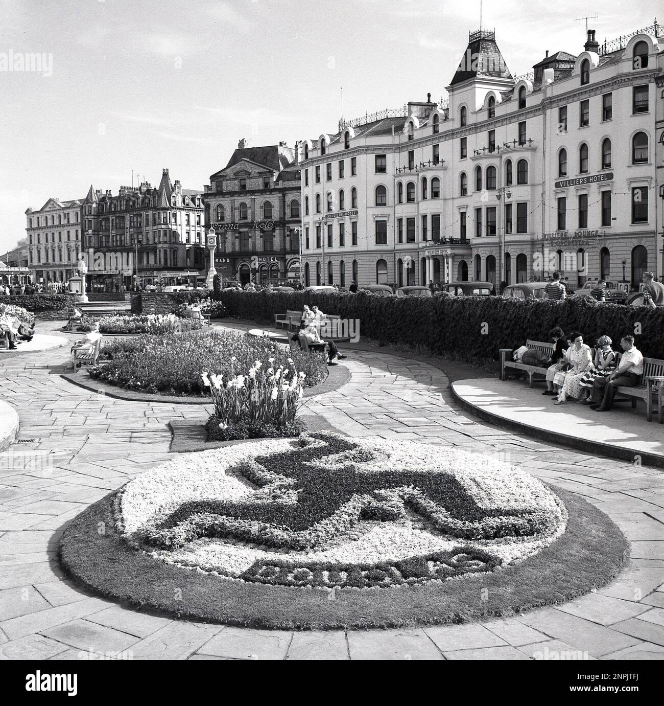 1950er Jahre, historisch, am Meer, Douglas, Isle of man, die Leute sitzen auf einer Bank in den Promenadengärten, die mit einem Manx-Blumensymbol dekoriert sind. Hotels des Tages sind zu sehen, darunter das Grand Hotel und das Villiers Hotel, mit einem Schild für die Lounge und das Orchester des Hotels. Stockfoto