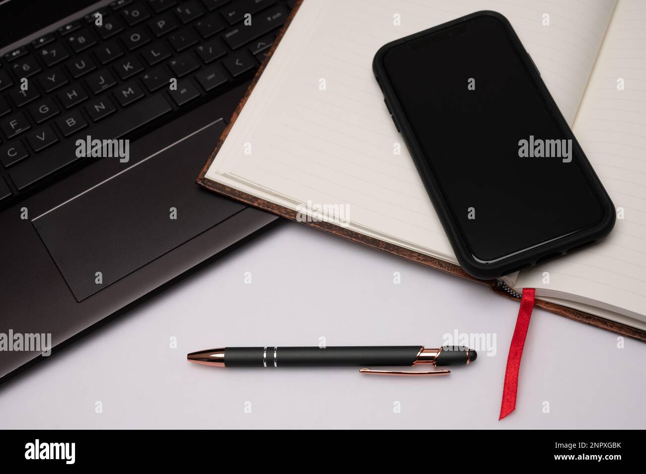 Ein mit Lesezeichen versehenes Notebook, Mobiltelefon, Stift und Laptop auf einem weißen Desktop-PC, das als Vorbereitung auf kreatives Schreiben das Gefühl eines Home Office vermittelt. Stockfoto