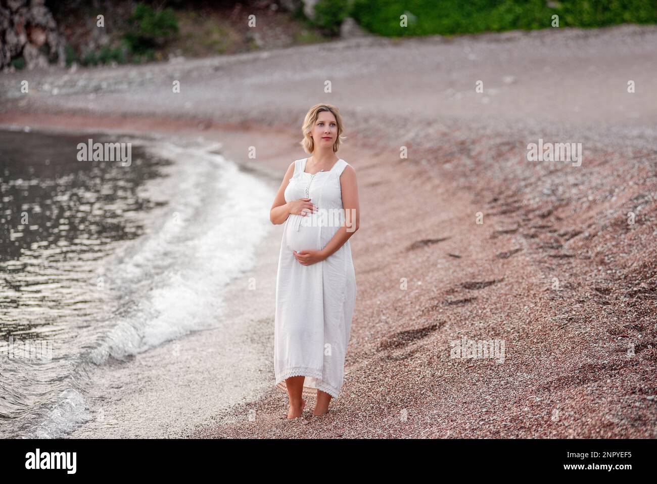 Porträt einer schwangeren Frau mittleren Alters in weißem Sonnenkleid. Werdende Mutter geht bei Sonnenuntergang am Meer entlang. Reisekonzept in der Schwangerschaft, gesund Stockfoto