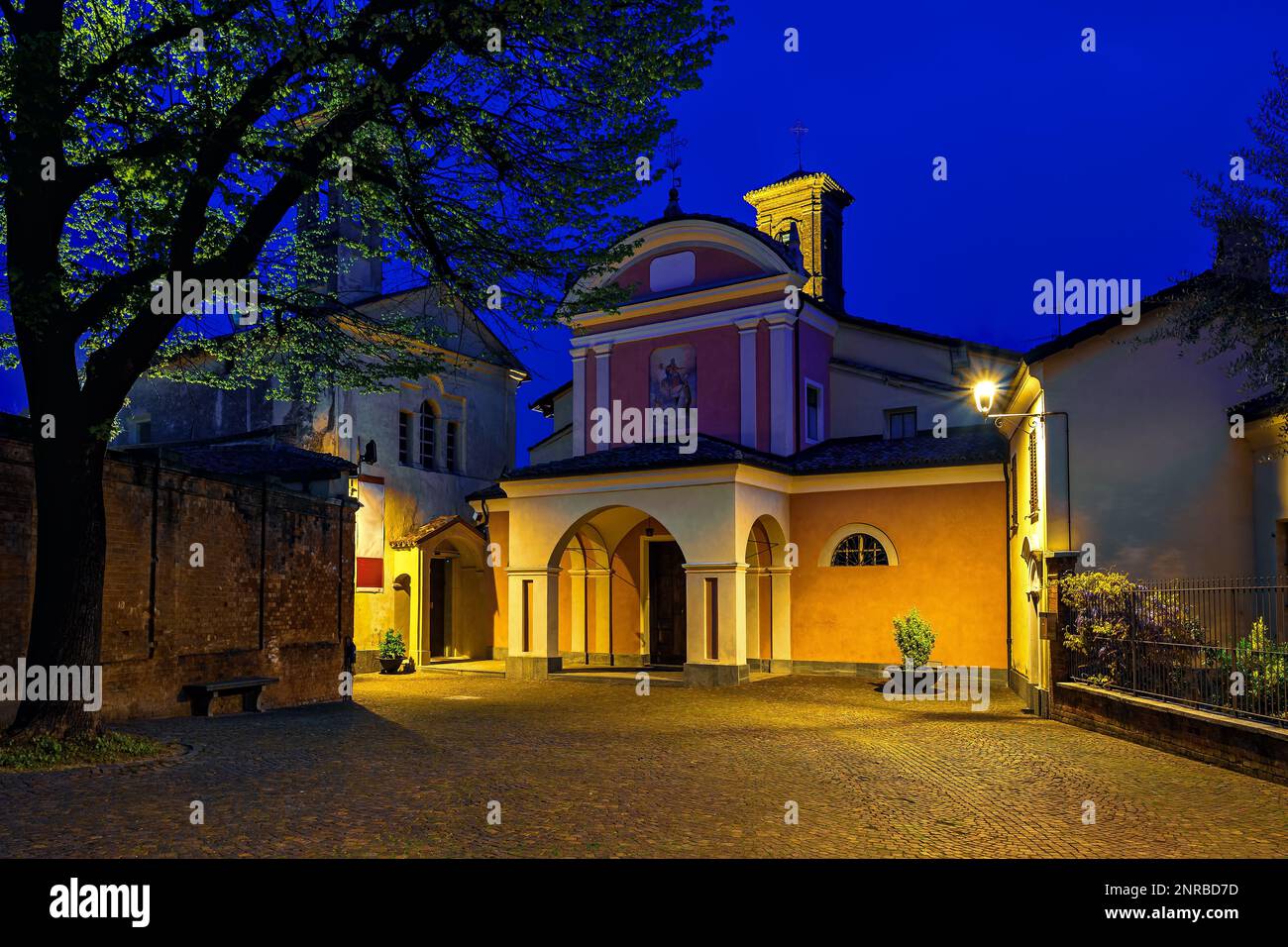 Beleuchtete Pfarrkirche auf einem kleinen Stadtplatz mit großem Baum unter blauem Himmel früh am Morgen in Barolo, Piemont, Norditalien. Stockfoto