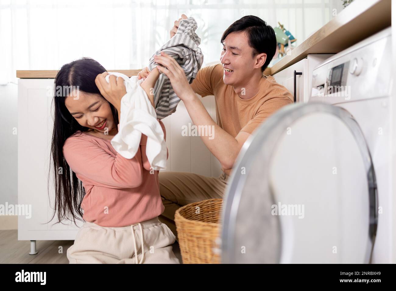 Junge Asiaten heirateten glückliches Paar lächelnd und machen Wäsche zu Hause. Freund und Freundin, die ihre Kleidung vor dem Laden der Waschmaschine zusammenlegen Stockfoto