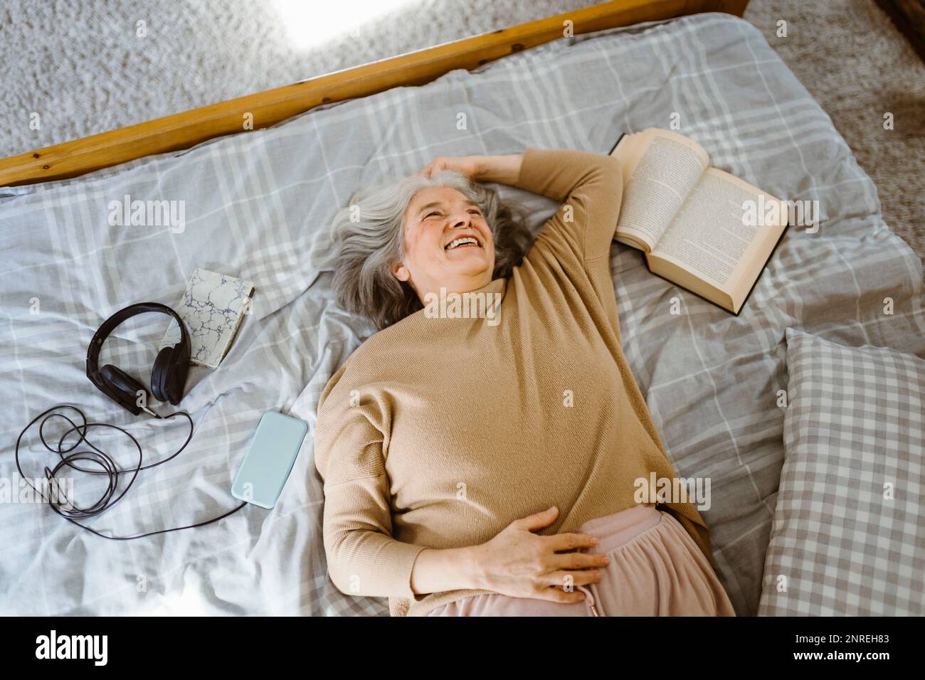 Glückliche Seniorin mit Buch und Kopfhörern, die auf dem Bett lagen Stockfoto