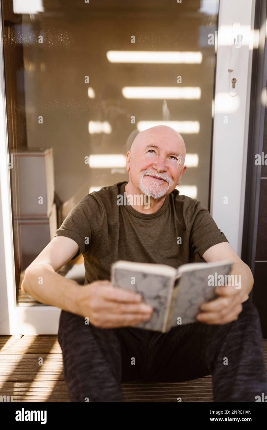 Lächelnder Seniorentag, träumte, während er zu Hause mit einem Buch saß Stockfoto