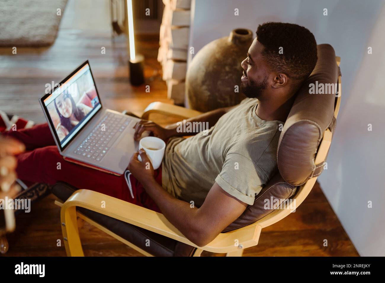 Lächelnder Mann, der mit einer Frau über einen Laptop Videoanrufe führt, während er zu Hause auf einem Stuhl sitzt Stockfoto