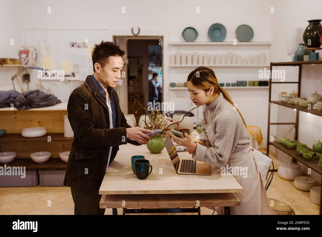 Männlicher Kunde mit Smartphone, der mit einer Töpferin spricht, während er in der Werkstatt bezahlt Stockfoto