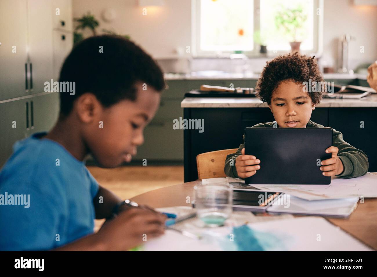 Ein Junge, der ein digitales Tablet benutzt, während der Bruder zu Hause ein Buch ausmalt Stockfoto