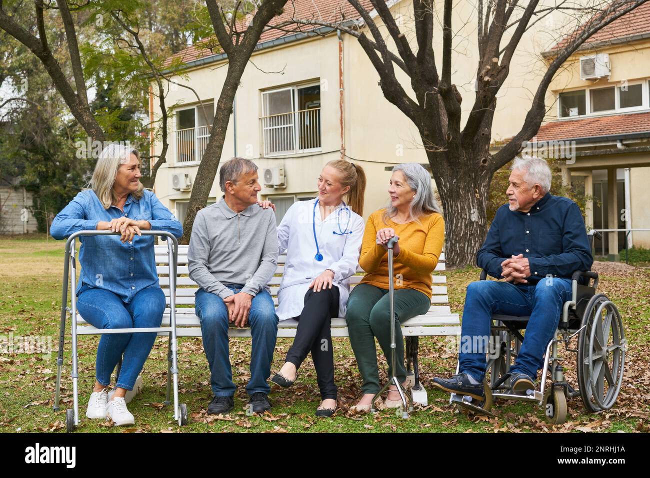 Arzt, der mit einer glücklichen Gruppe älterer Menschen spricht, die auf einer Bank im Garten des Pflegeheims sitzt Stockfoto