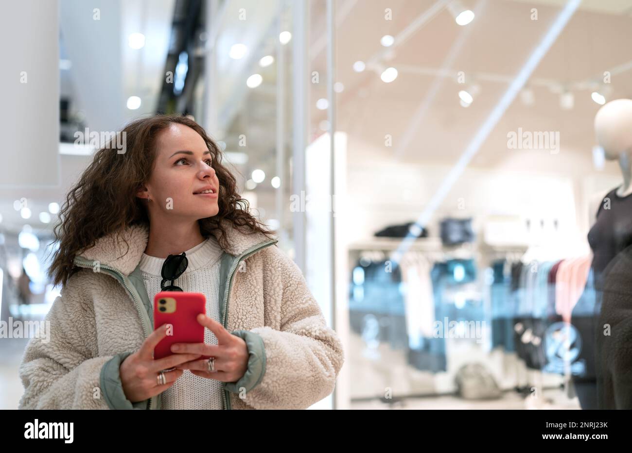 Eine junge Frau, die durch das Einkaufszentrum läuft, auf ein Fenster von Modegeschäften schaut und ihr Handy in der Hand hält. Stockfoto