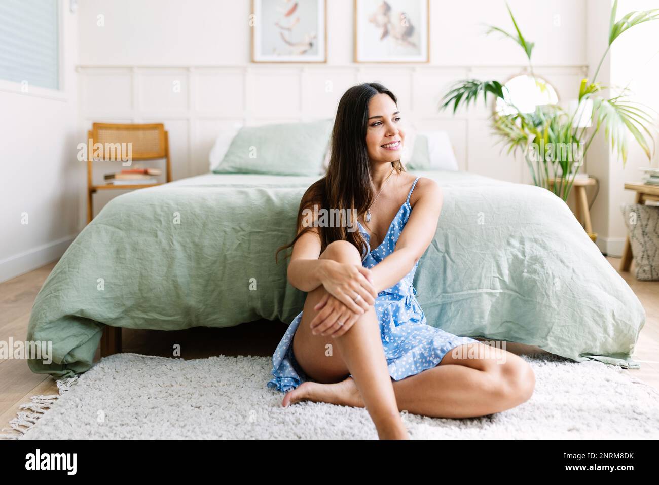Fröhlich entspannte junge Erwachsene Frau, die auf dem Teppich im Schlafzimmer saß und wegschaute Stockfoto