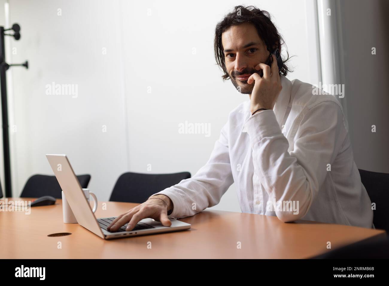 Porträt eines jungen Geschäftsmanns oder Büromitarbeiters, der mit seinem Laptop in einem gemütlichen Büro arbeitet und mit seinem Mobiltelefon kommuniziert Stockfoto