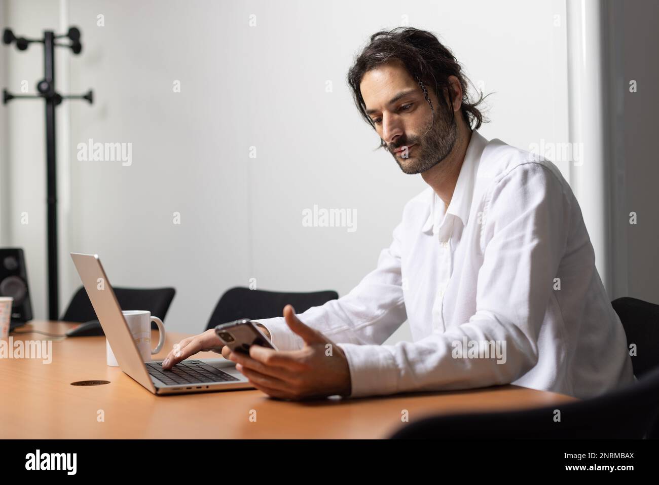Porträt eines Mannes, der an seinem Laptop arbeitet und mit seinem Telefon kommuniziert. Er sitzt in einem schönen Büro Stockfoto