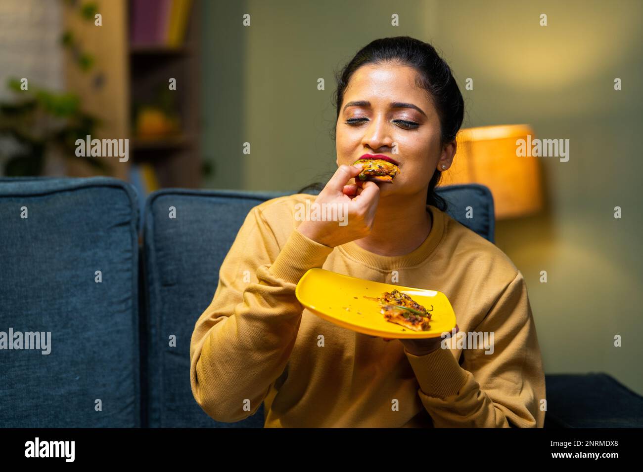 Ein mittelgroßes Mädchen isst gerne leckere Pizza, indem es die Augen zu Hause auf dem Sofa schließt – Konzept von leckerem Essen, Aufregung und Emotionen oder Ausdruck. Stockfoto