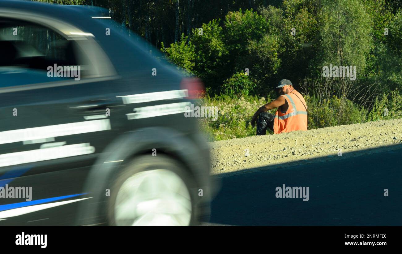 Ein Straßenarbeiter in einer orangefarbenen Weste sitzt am Straßenrand vorbei an vorbeifahrenden Autos und entspannt sich im Sommer in Russland. Stockfoto
