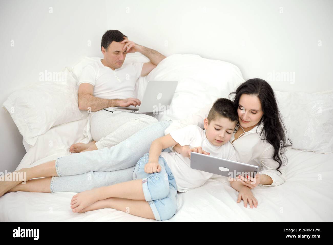Jeder hat seinen eigenen Job, Vater arbeitet zu Hause mit einem Laptop. Die mutter verdient Geld. sie unterhält den Sohn mit einem Tablet, weiße Kleidung, ein Bett, Ruhewohnungen verbringen Zeit zusammen und sind allein mit der Familie Stockfoto
