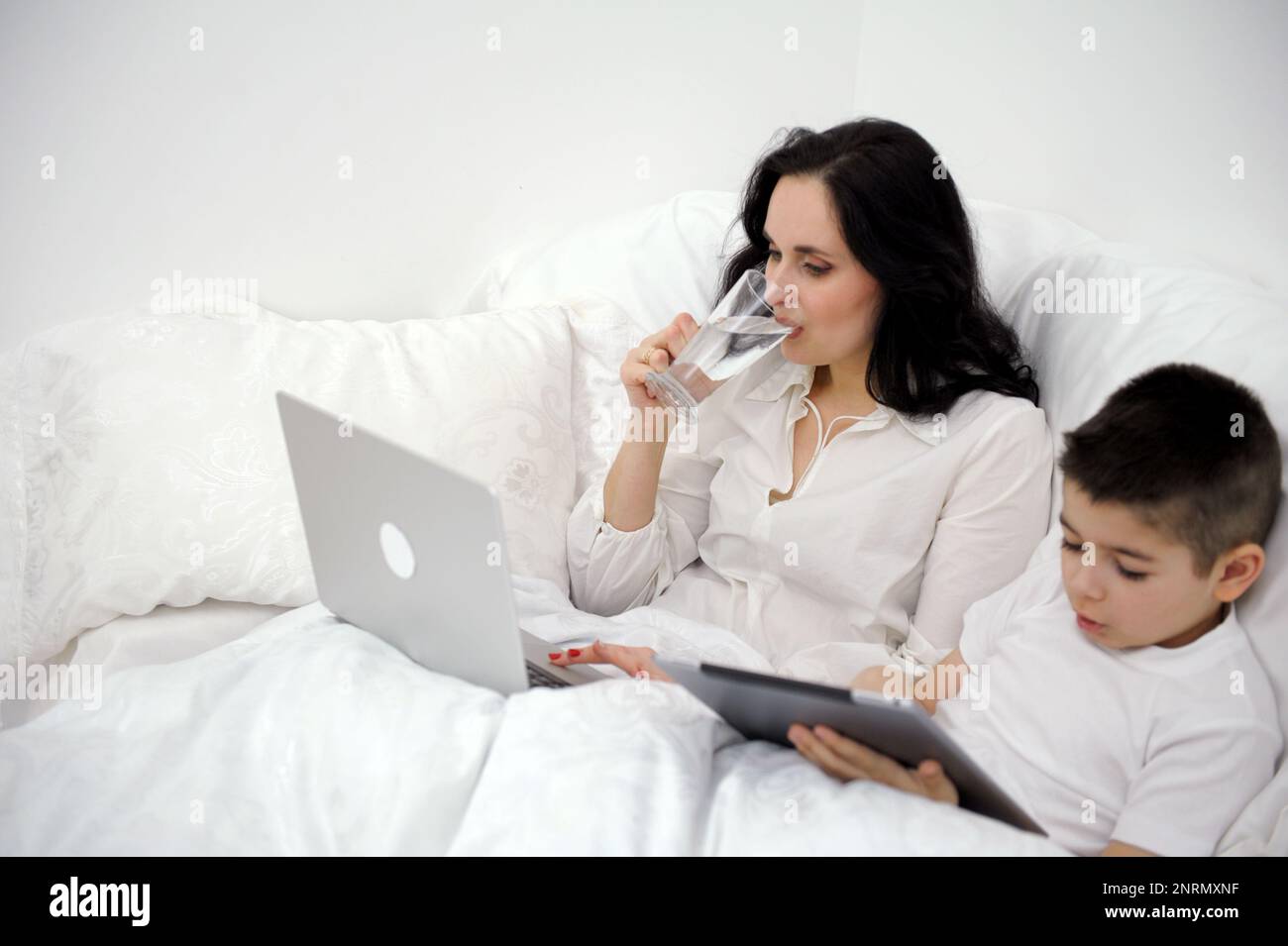 Diät Gewichtsverlust Frau trinkt Wasser im Bett während der Arbeit auf Computer Sohn spielt auf Tablet richtige Ernährung gesunde Lebensweise krank zu Hause Coronavirus Quarantäne Ruhe nach der Arbeit verbringen Zeit Stockfoto