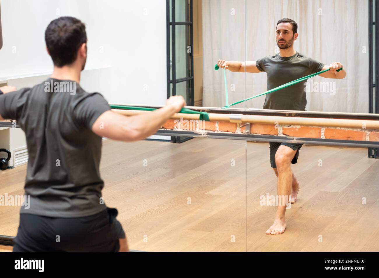 Rückansicht des konzentrierten jungen bärtigen männlichen Athleten in Sportswear, der das Widerstandsband zieht, während er vor dem Spiegel die Schulterstabilität trainiert Stockfoto