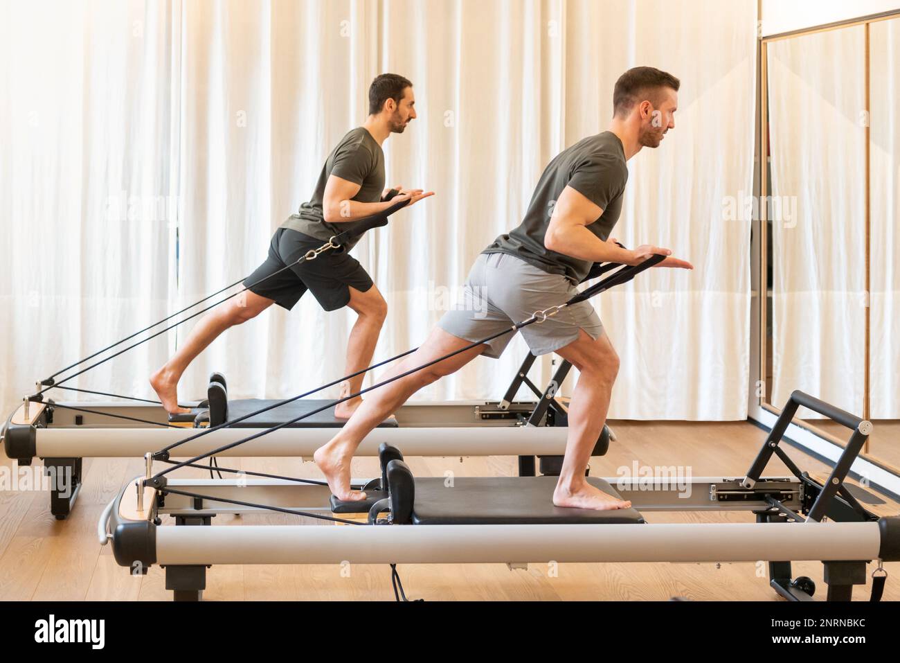 Ganzkörper-Seitenansicht von konzentrierten männlichen Sportlern in aktiver Kleidung, die Stehbaumumarmungen Pilates Training auf professionellen Geräten im Fitnessstudio Stockfoto