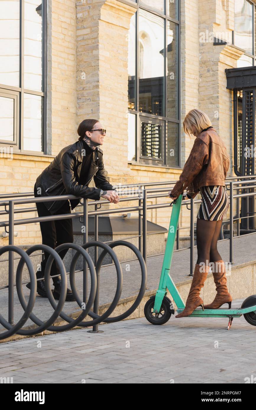 Blonde Frau in stilvollem Outfit und Stiefel auf Absatz stehen auf einem Elektroroller neben einem fröhlichen tätowierten Mann, Stock Image Stockfoto