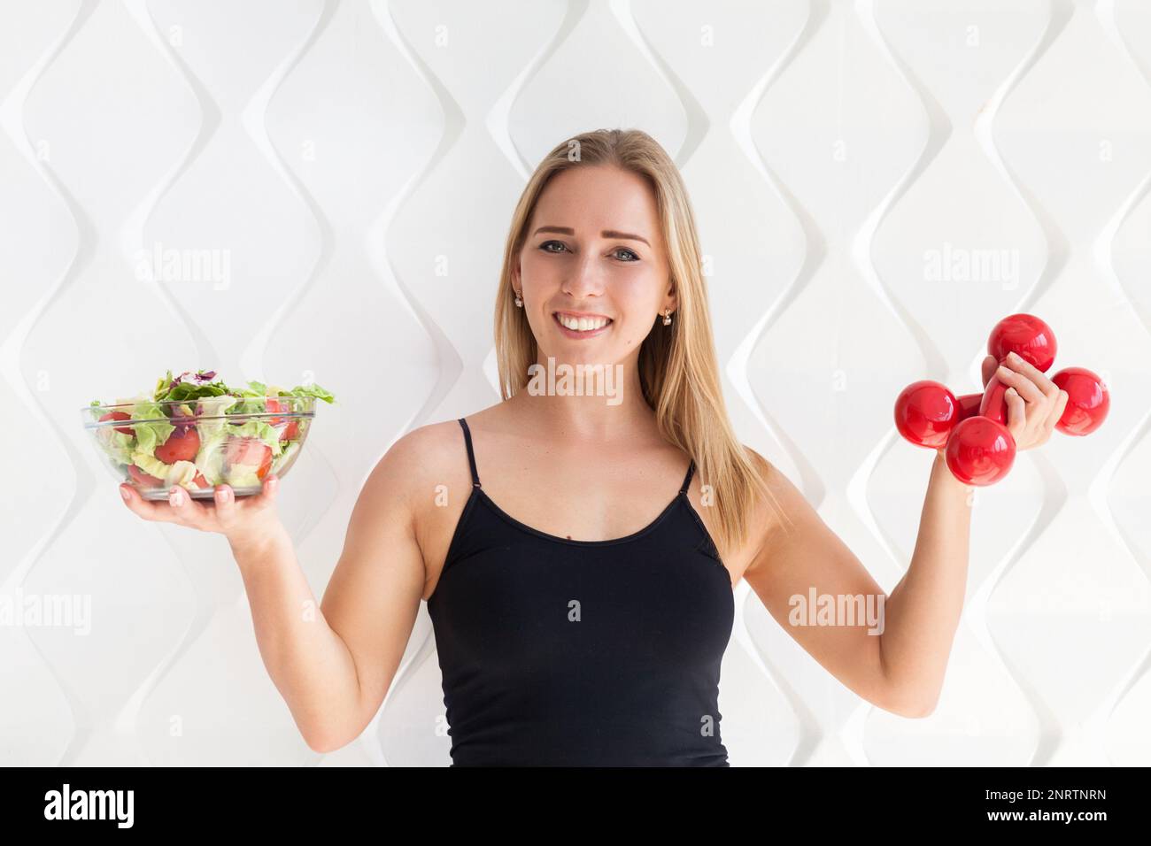 Junge Sportlerin in Sportbekleidung mit roten Kurzhanteln und grünem Salat in Glasschale. Gesundes Leben Bodybuilding Lifestyle Konzept Bild, Stockfoto