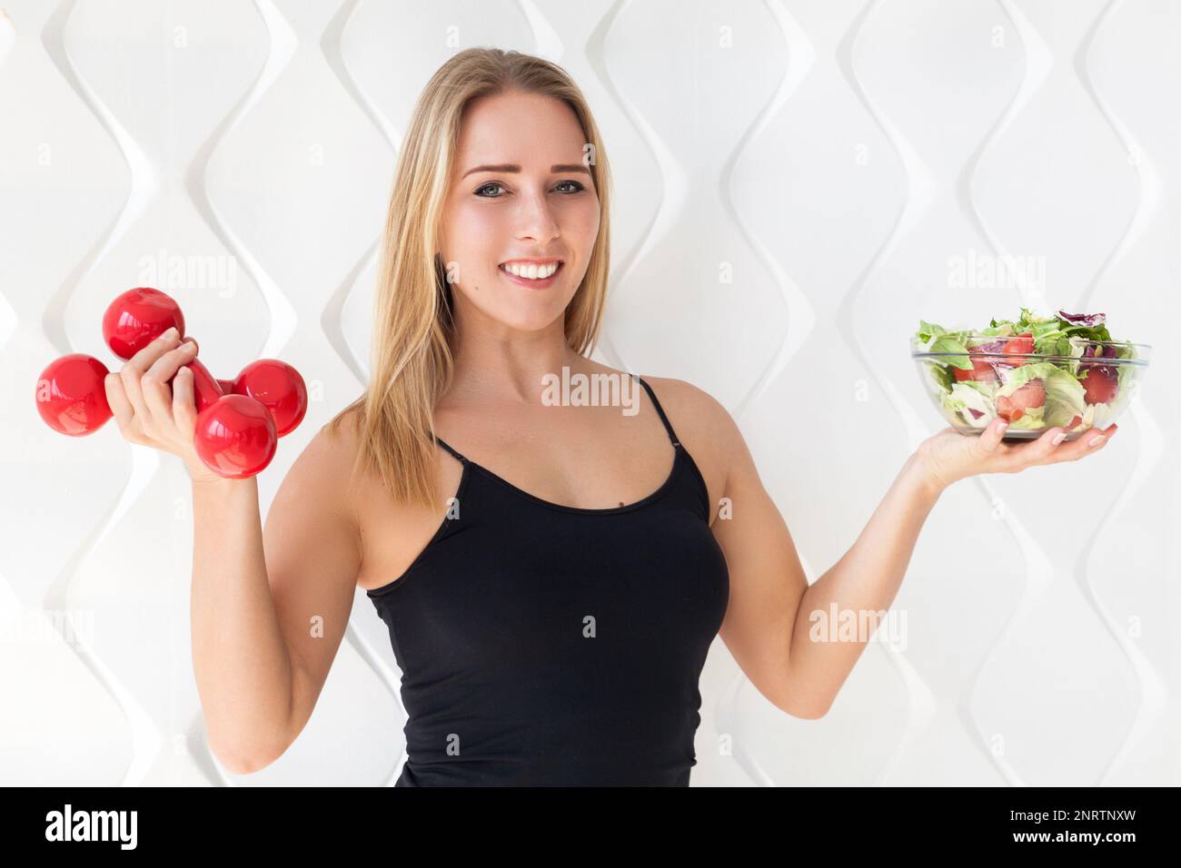 Junge Sportlerin in Sportbekleidung mit roten Kurzhanteln und grünem Salat in Glasschale. Gesundes Leben Bodybuilding Lifestyle Konzept Bild, Stockfoto