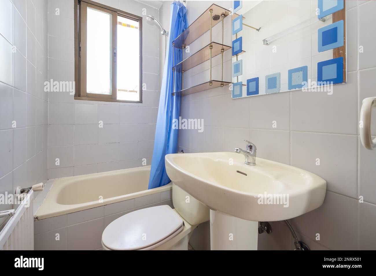 Kleines Badezimmer mit Aluminiumfenstern, einem rahmenlosen Spiegel mit blauen Mustern, einem Porzellanwaschbecken mit einem Porzellansockel und einer kleinen Badewanne mit einem Schlafzimmer Stockfoto