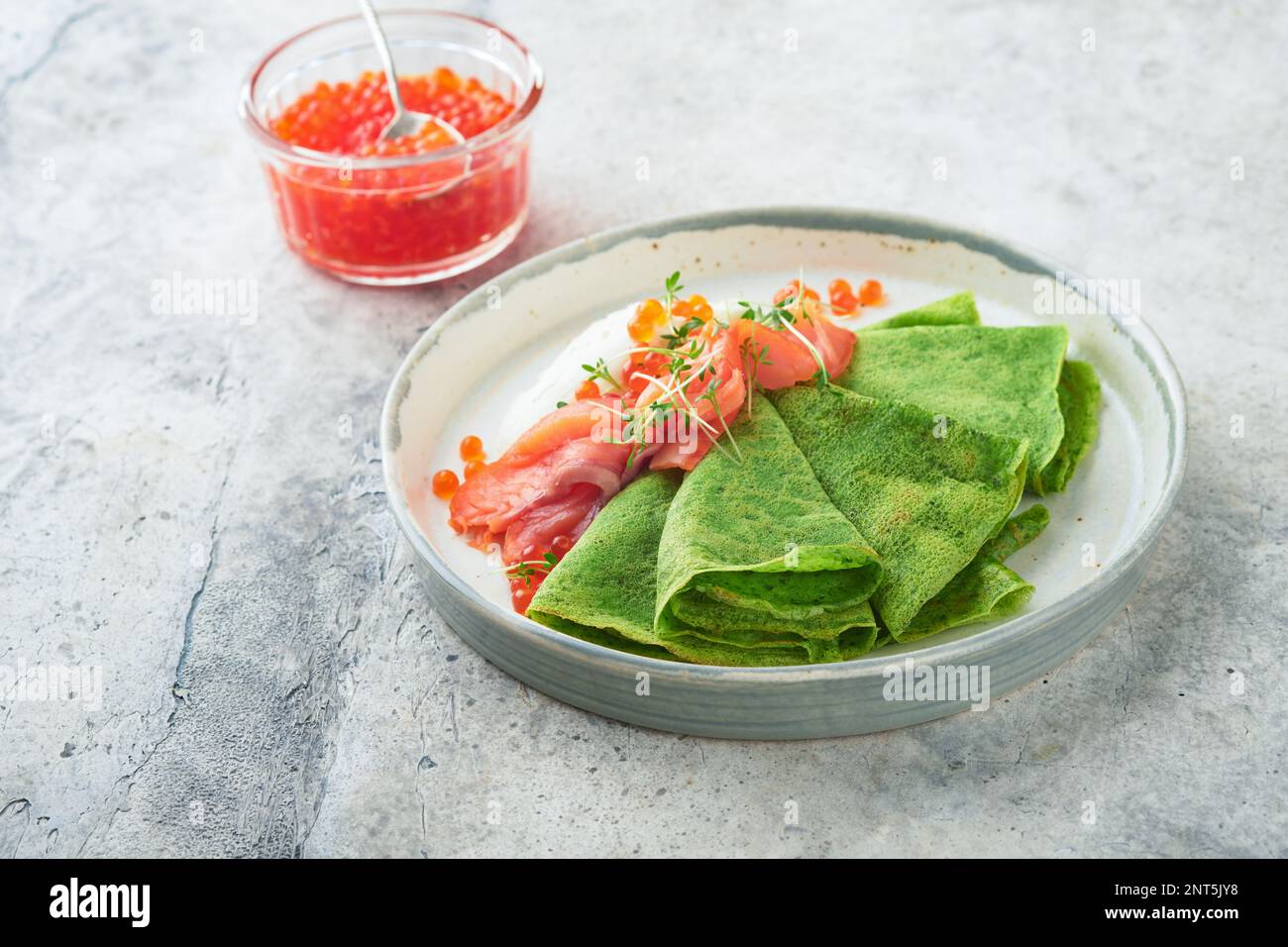 Grüne Pfannkuchen. Runde, dünne, grüne Spinat-Crepe-Pfannkuchen mit rotem und schwarzem Kaviar und Joghurt-Sauce auf grauem Betonhintergrund. Gesundes veganes Frühstück Stockfoto