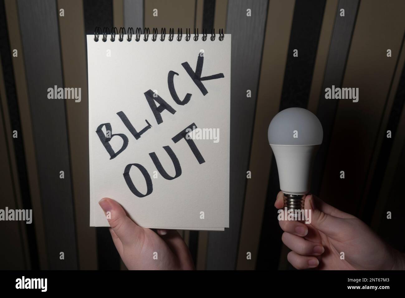 Ein Notizblock mit der Aufschrift „Blackout“ in der einen Hand und eine elektrische Glühbirne in der anderen Hand (Nahaufnahme). Stromausfallkonzept. Energiekrise Stockfoto