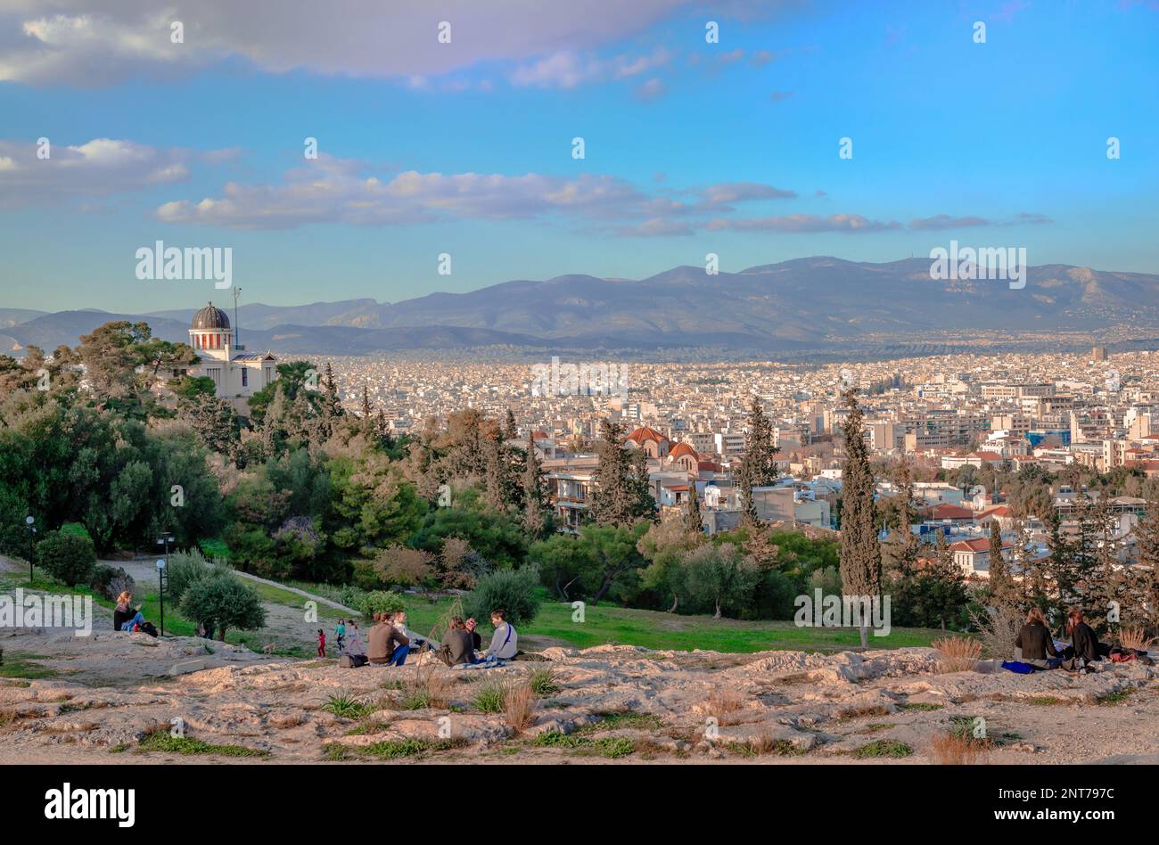 Zufällige Menschen auf dem Hügel von Pnyx genießen die Skyline von Athen, Griechenland an einem sonnigen Nachmittag im Winter. Das Nationale Observatorium befindet sich auf der linken Seite. Stockfoto