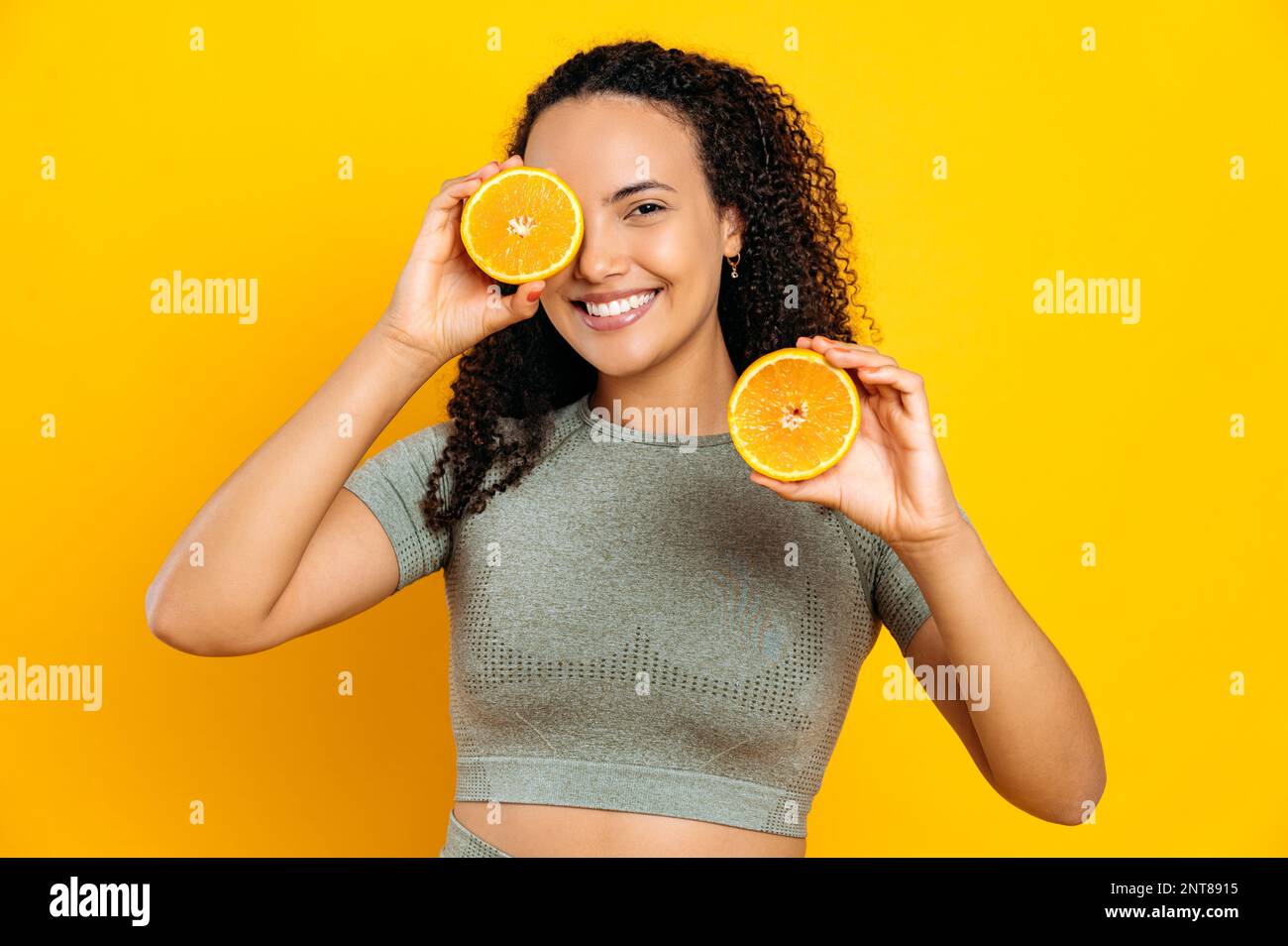 Konzept eines gesunden Lebensstils. Glückliche, reizende brasilianische oder hispanische junge, lockige Frau, in Sportkleidung, steht auf isoliertem gelben Hintergrund, hält zwei Hälften orange, bedeckt ein Auge, lächelt Stockfoto