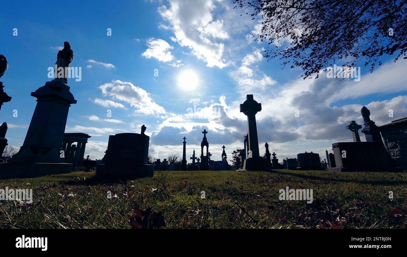 Schattige Grabsteine auf dem Friedhof mit Silhouetten und überqueren niedrige Winkel an einem sonnigen Himmel mit wackelnden Wolken Stockfoto