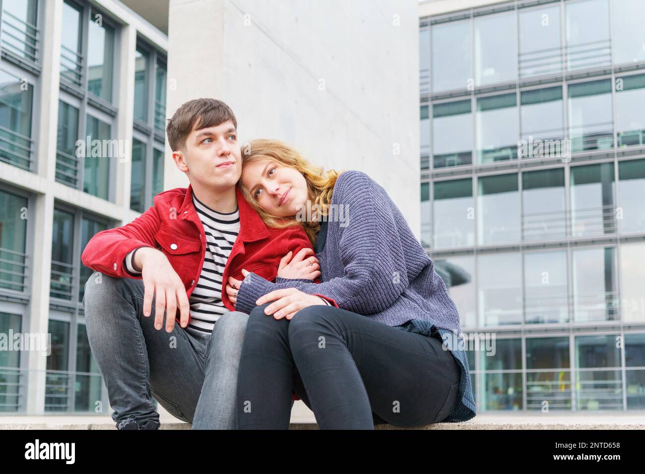 Ein junges, liebevolles, heterosexuelles Paar sitzt auf einer Treppe vor einem modernen Gebäude mit Glasfassade Stockfoto