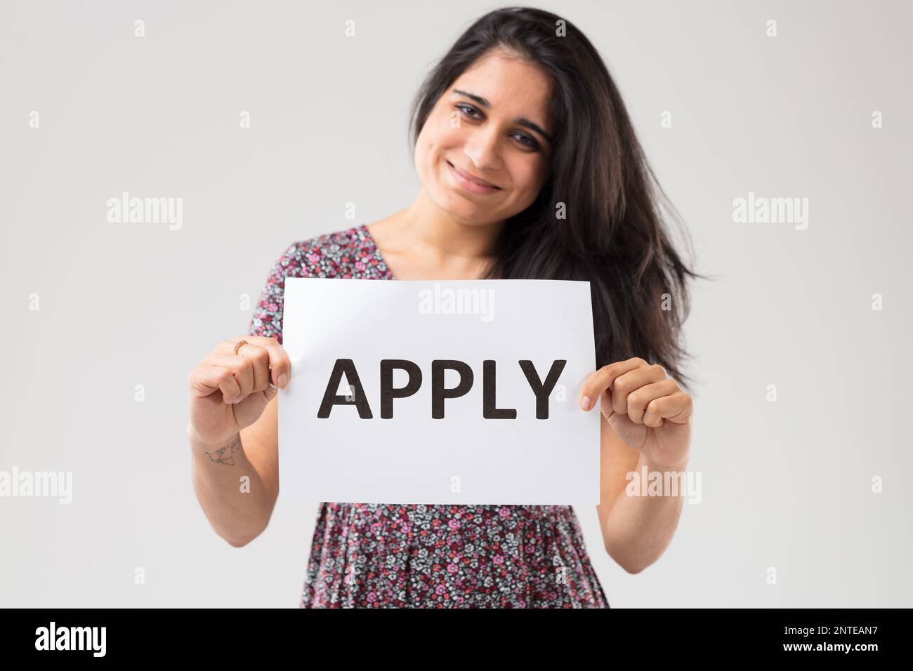 Porträt eines indischen Mädchens in einem blumigen Kleid und langen Haaren mit einem Schild. Sie ist glücklich und lächelt und auf dem Schild steht "Bewerben", was dich einlädt, weil ich es bin Stockfoto