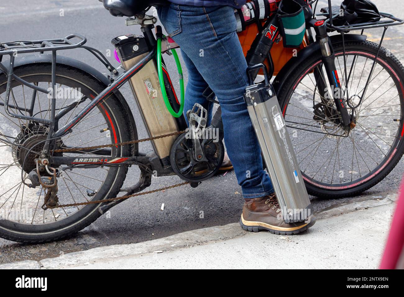 Lithium-Ionen-E-Bike-Batterien, die von einem Lieferboten getragen werden; eine auf dem E-Bike und eine Ersatzbatterie an seinem Fuß. Stockfoto