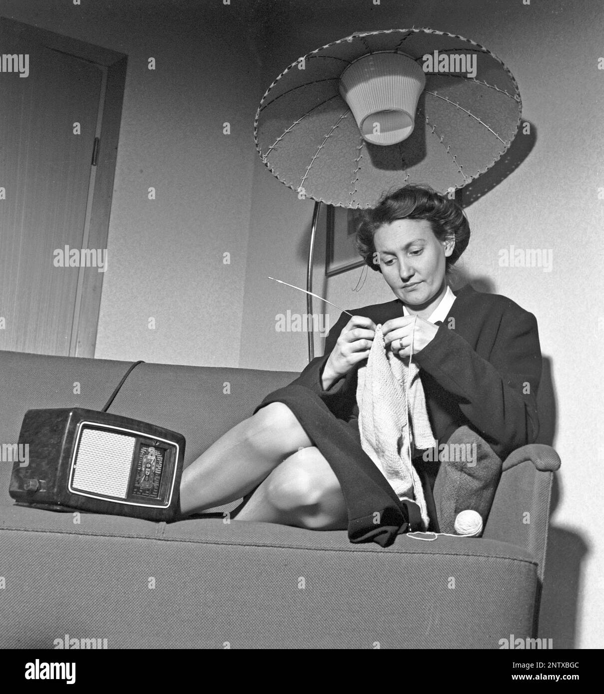 Radio-Hörer in der Vergangenheit. Eine Frau sitzt auf einer Couch und strickt mit einem Radio an der Seite. Auf der Vorderseite des Radios befindet sich eine Waage, und als Sie das Wahlrad daneben gedreht haben, sodass sich der Zeiger in der Mitte eines Sendernamens befand, war dieser Sender dort. Neben UKW-Übertragungen hörten sie auch ausländisches Radio auf Langwellen. Schweden 1945 Kristofferson Ref. L179-3 Stockfoto