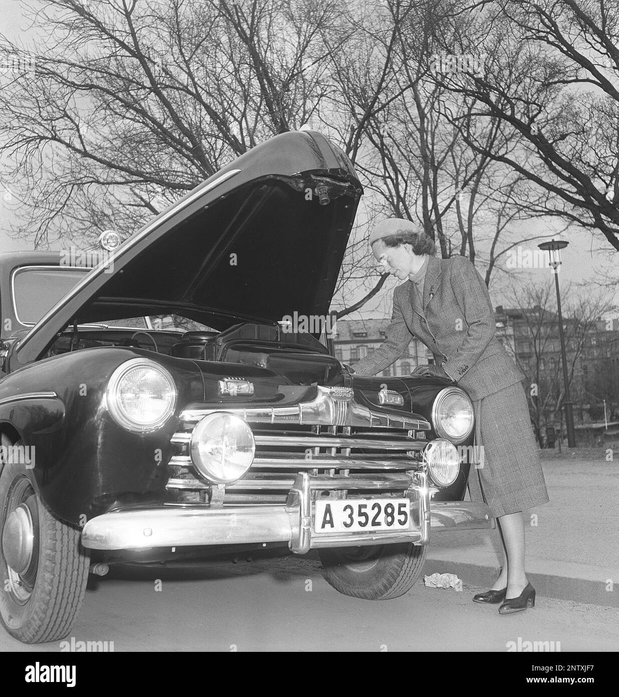 In den 1940er Jahren. Eine hübsch gekleidete Frau mit passender Jacke und passendem Rock hat die Motorhaube ihres Ford-Autos geöffnet und schaut sich etwas an. Nach ihrer Kleidung zu urteilen, ist sie nicht gekleidet, um mit mechanischen Problemen umzugehen, noch irgendwelche Werkzeuge oder Flüssigkeiten sichtbar, nur ein Foto, das zu diesem Zeitpunkt eine Do-it-yourself-Frau darstellt. Schweden 1949Kristoffersson Ref. AY83-10 Stockfoto