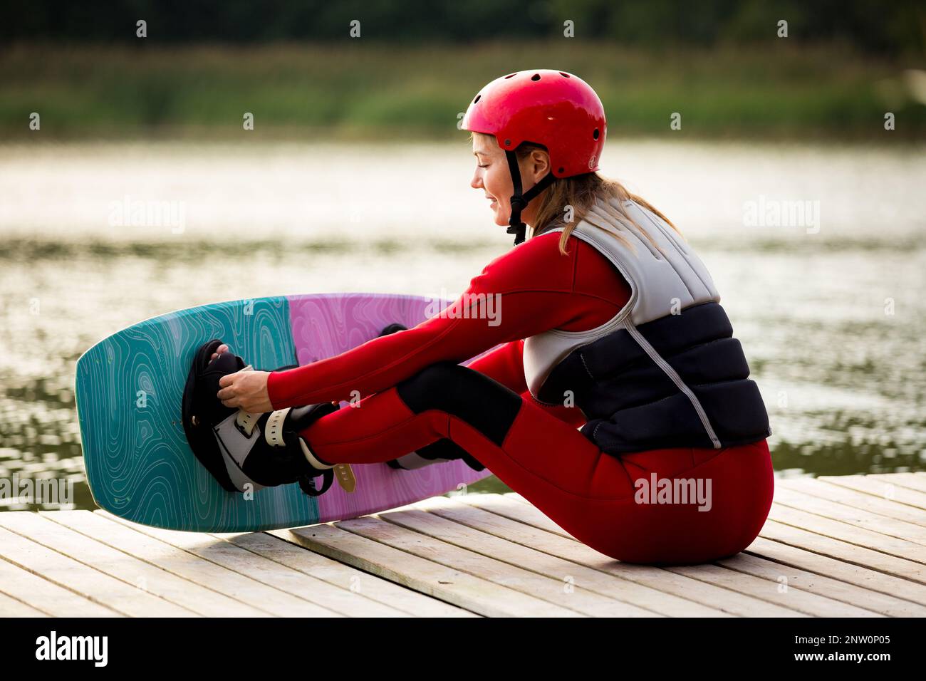 Frau in Neoprenanzug, Helm und Schwimmweste, die mit Wakeboard auf einem hölzernen Pier sitzt. Sonniger Sommertag. Sicherheit im Sport. Wassersport in Finnland. Versicherungen Stockfoto