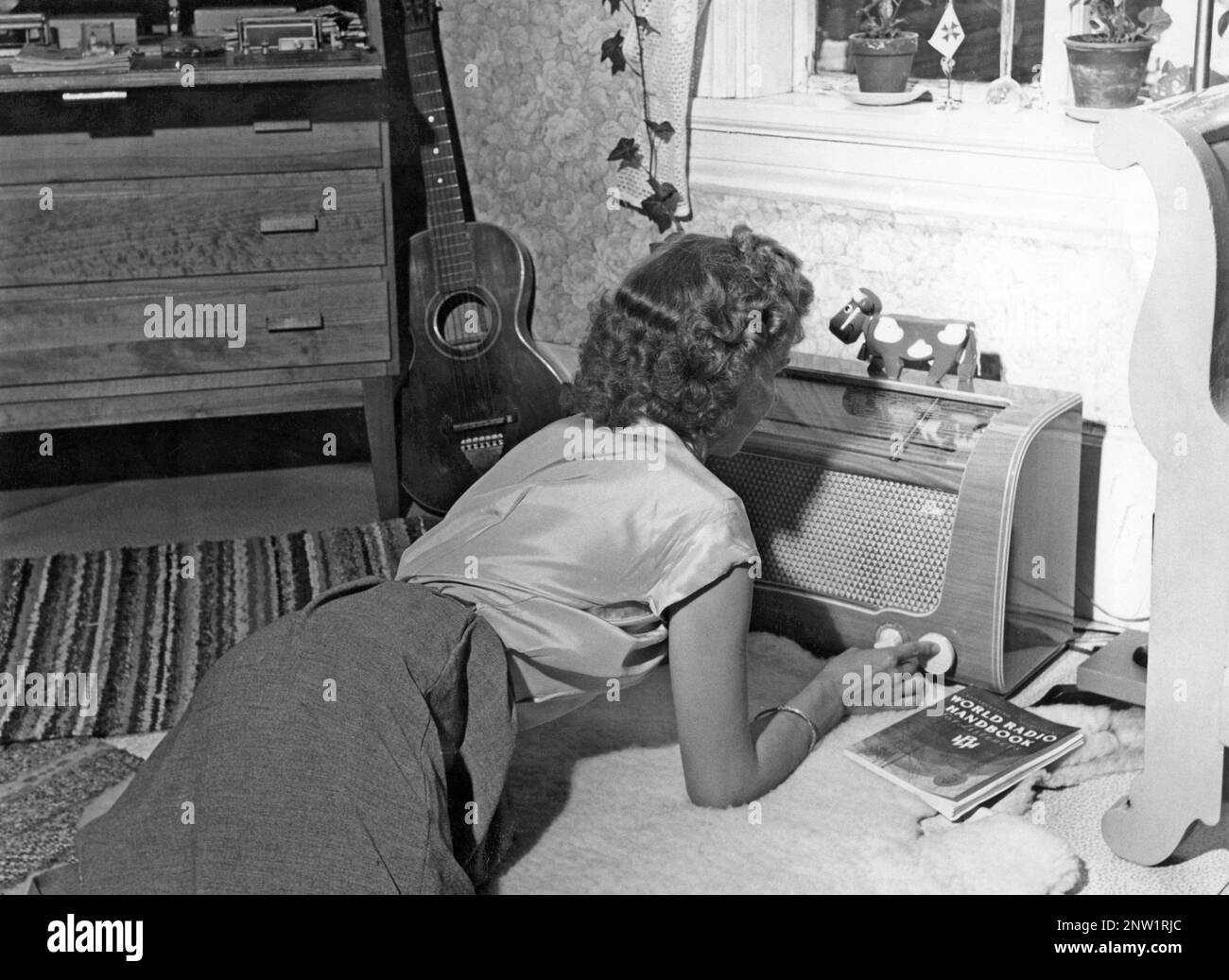 Die junge Kurzwellenfreundin Gertrud Larsson zu Hause bei ihrem Radio. Auf der Vorderseite des Radios befindet sich eine Waage, und als Sie das Wahlrad daneben gedreht haben, sodass sich der Zeiger in der Mitte eines Sendernamens befand, war dieser Sender dort. Neben UKW-Übertragungen hörten sie auch fremde Radiosender auf Langwellen- und Kurzwellen. Neben ihr steht der klassische Katalog World Radio Handbuch, in dem alle internationalen Radiosender aufgeführt sind und auf welchen Frequenzen sie übertragen werden. Schweden 1953 Stockfoto