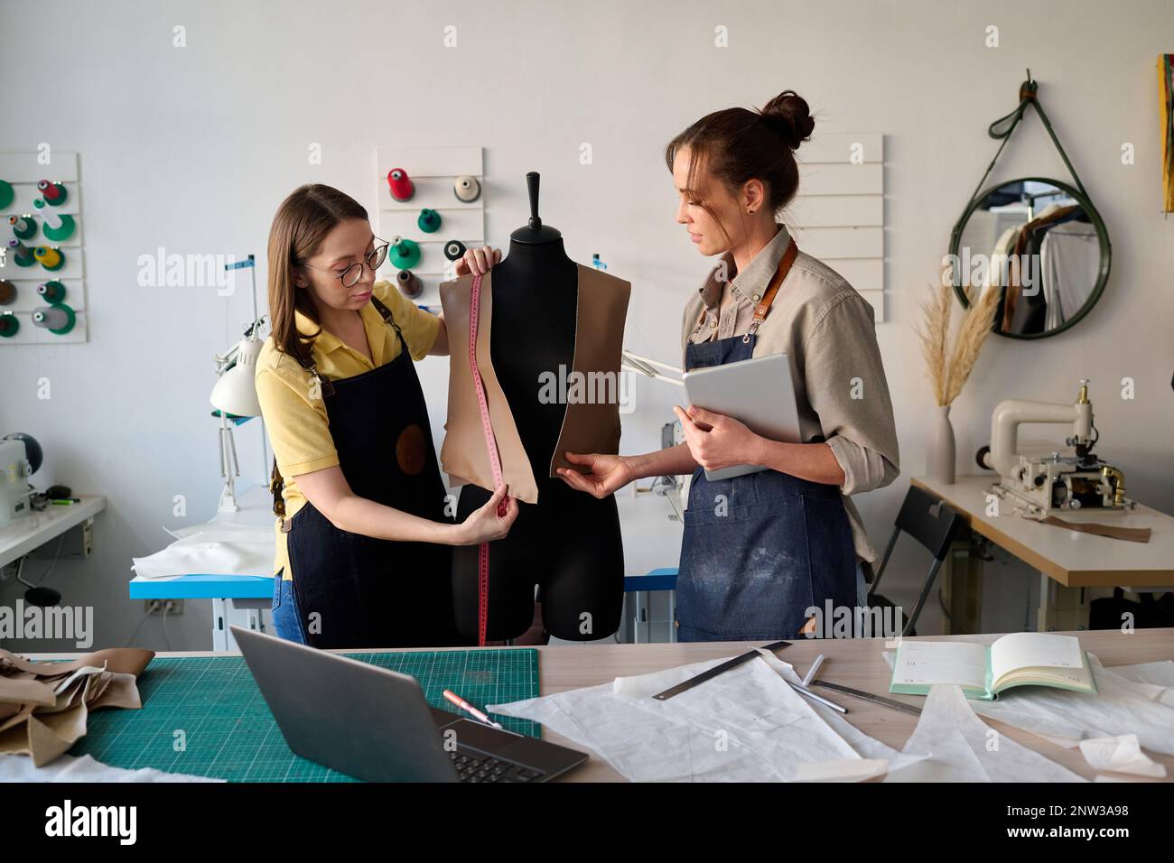 Junger Angestellter des Schneider, der ihr dabei hilft, Maßnahmen in Bezug auf eine unfertige Lederweste zu ergreifen, während beide vor der Schaufensterpuppe stehen Stockfoto