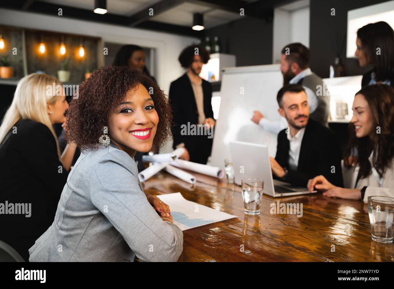 Porträt einer jungen lächelnden lateinischen eleganten Frau, die während eines Meetingbüros am Schreibtisch sitzt. Kollegen, die im Reden arbeiten. Stockfoto