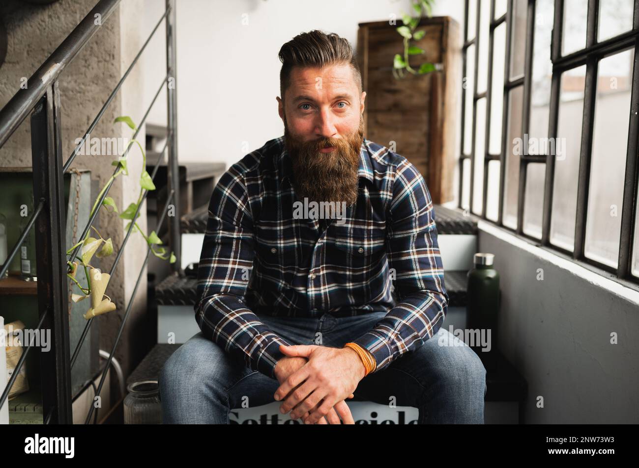 Porträt eines Hipstermanns, der in die Kamera schaut. Ein Kerl, der auf der Treppe sitzt, mit selbstbewusstem Gesicht. Stockfoto