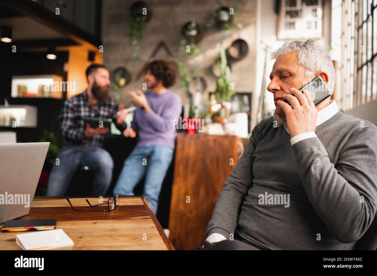 Ein Geschäftsmann, der am Telefon telefoniert, sitzt am Schreibtisch in einem modernen Büro, während Kollegen über Hintergrundgespräche diskutieren. Stockfoto