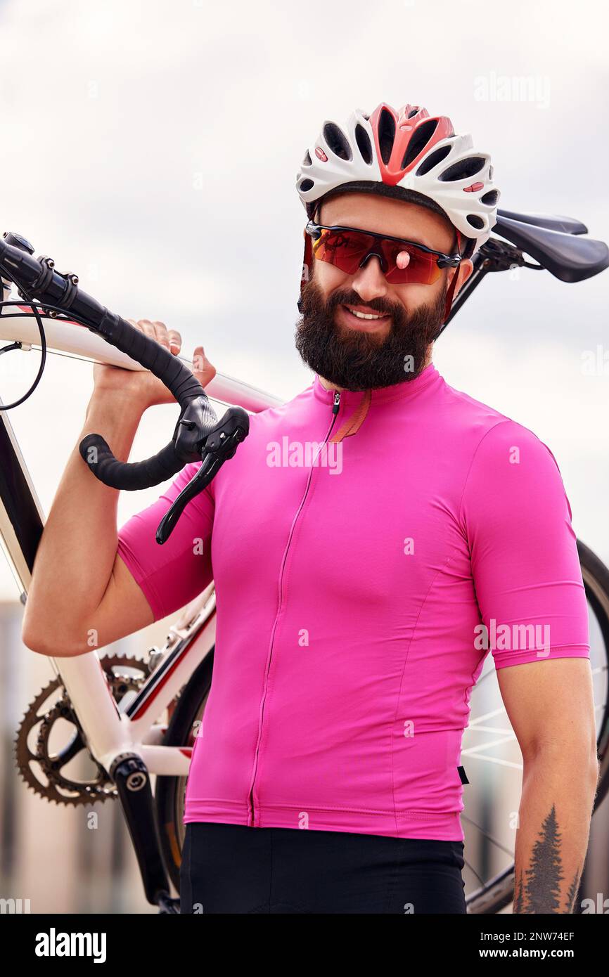 Bärtiger männlicher Radfahrer mit Brille und ein Helm schaut auf die Kamera mit einem Fahrrad in der Hand gegen den blauen Himmel, Lifestyle, Cardio-Training Stockfoto