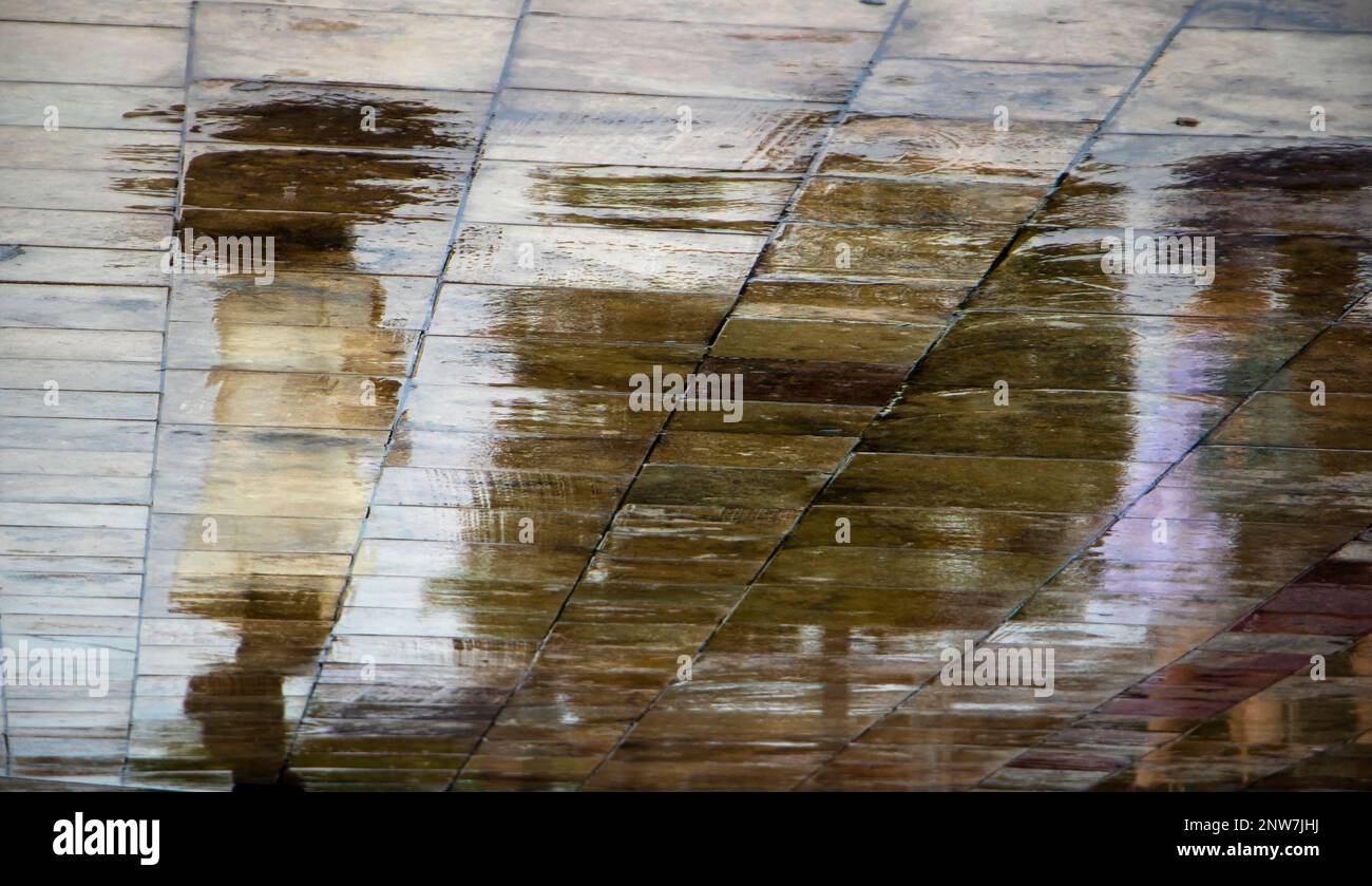 Abstrakte, verschwommene Silhouettenschattenreflexionen einer nicht wiedererkennbaren Person im weißen Trenchcoat, die unter dem Schirm auf nassen Straßenpflastern einer Stadt auf einer Ra läuft Stockfoto