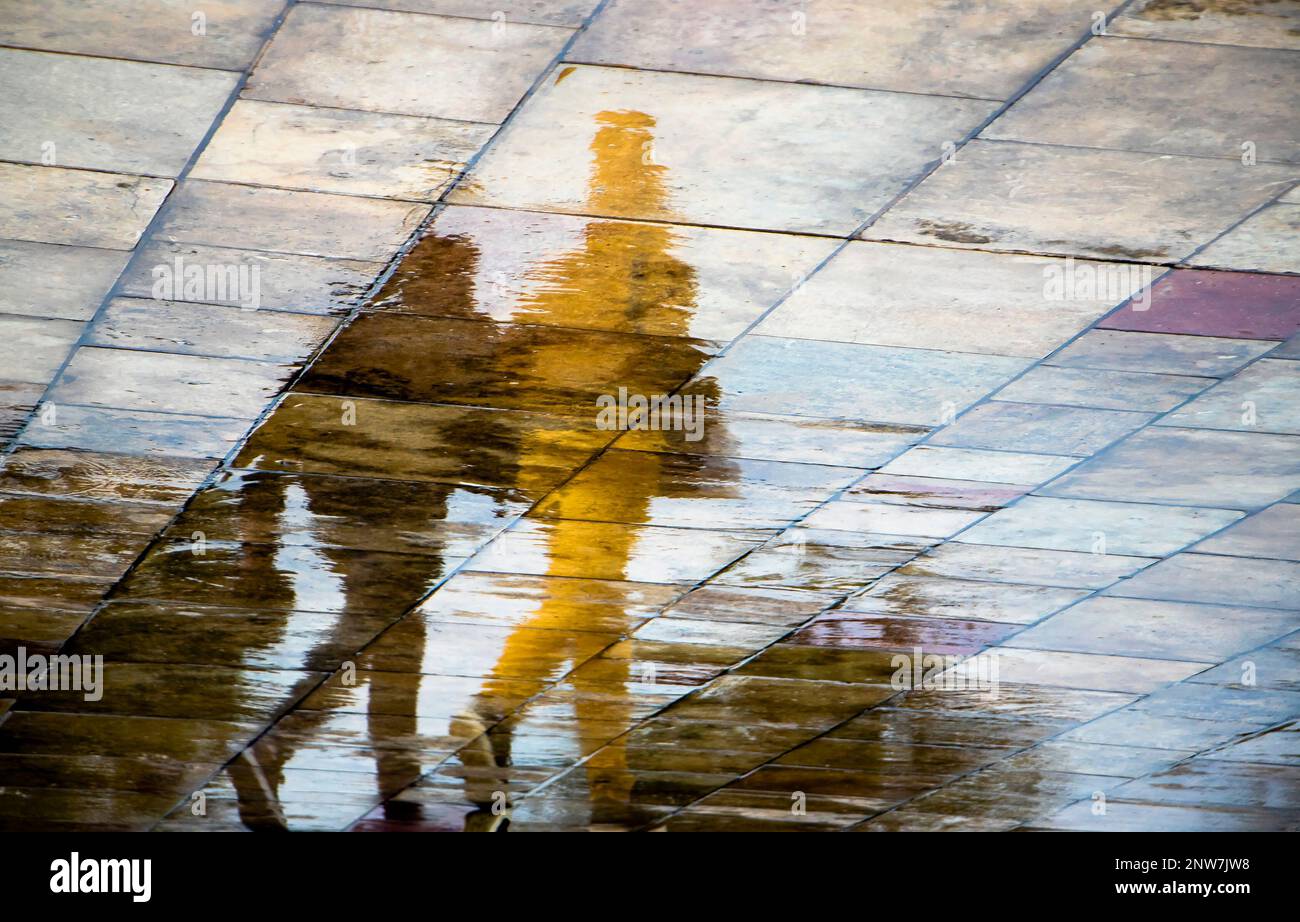 Abstrakte, verschwommene Silhouettenschattenreflexionen von nicht wiedererkennbaren Menschen, die an regnerischen Tagen unter einem Schirm auf nassen Straßenbelägen in der Stadt spazieren Stockfoto