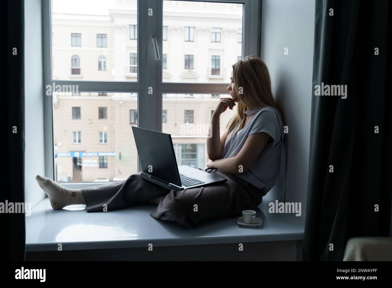 Eine junge Frau sitzt auf einer Fensterbank, arbeitet an einem Laptop, schaut aus dem Fenster. Geschäfts- und Bildungskonzept. Stockfoto