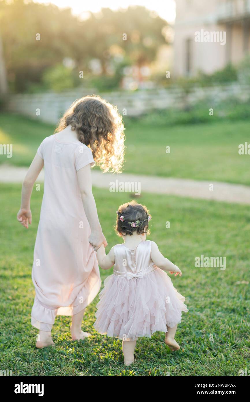 Die große Schwester hilft der kleinen Schwester, mit schwacher Sonne in blassrosa Kleidern und Blumenkronen zu gehen Stockfoto